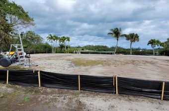 Cleared land with palm trees under a cloudy sky, bordered by a black silt fence.