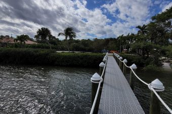 A wooden boardwalk extends over water under a cloudy sky with palm trees nearby.