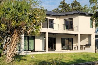 Modern two-story house with large windows and balcony surrounded by greenery.