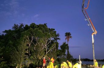 Workers pour concrete at dusk on a construction site surrounded by trees.