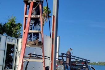 Man on ladder repairing electrical pole near river under clear sky.