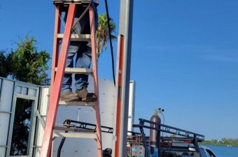 Man on ladder repairing electrical pole near river under clear sky.