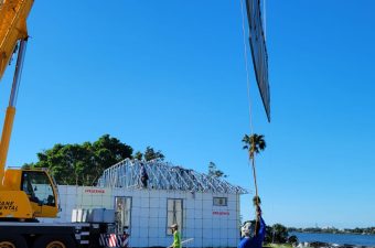 Workers use cranes to lift a large metal structure beside a waterfront.