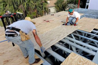 Two workers installing plywood on a roof frame outdoors.