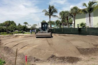 A bulldozer levels soil at a construction site with palm trees in the background.