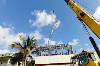Crane lifting a metal structure against a blue sky with palm trees nearby.