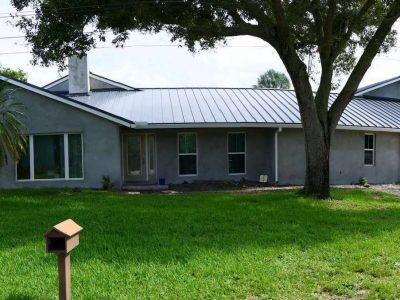 A single-story house with a metal roof and a large tree in the front yard.