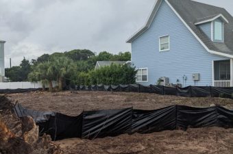 A residential construction site with soil excavation and a blue house in the background.