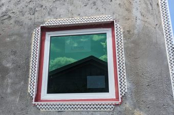 Square window on textured gray wall reflecting a rooftop and sky.