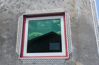 Square window on textured gray wall reflecting a rooftop and sky.